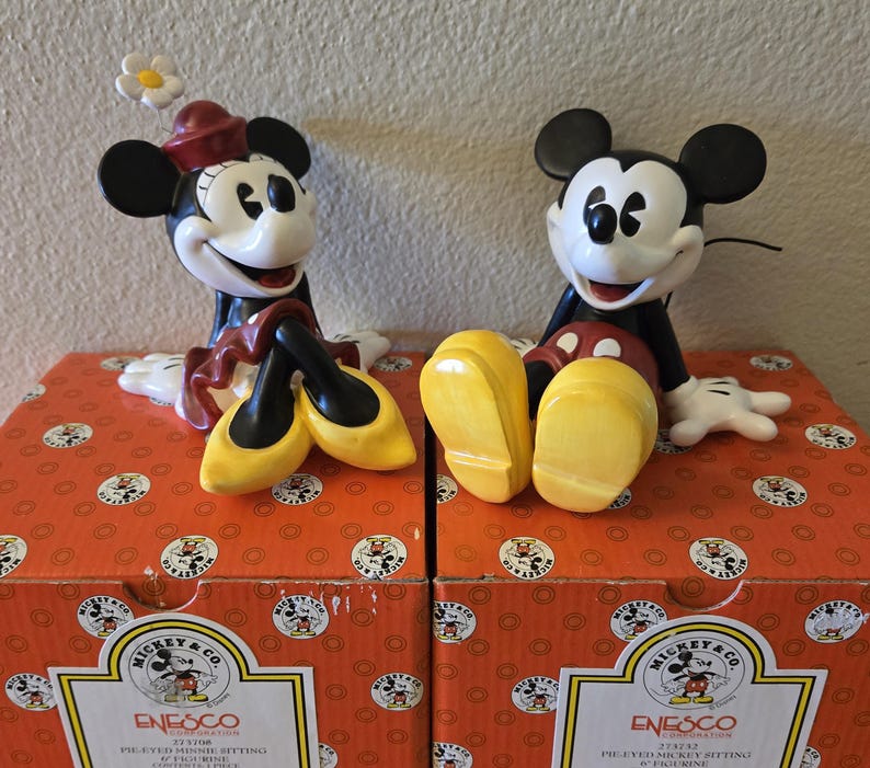 Puede incluir: Dos figuras de cer&aacute;mica de Mickey y Minnie Mouse sentadas en cajas. Minnie lleva un sombrero rojo y zapatos amarillos, y Mickey tiene zapatos amarillos. Las cajas son naranjas con dise&ntilde;os de Mickey Mouse y el logo de Enesco.