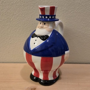May include: A ceramic pitcher shaped like Uncle Sam, featuring a red, white, and blue color scheme. The pitcher has a handle and is decorated with a top hat, bow tie, and striped pants. The pitcher is approximately 10 inches tall.
