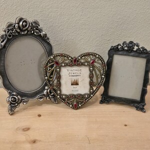 May include: Three decorative silver-toned picture frames. The frames have ornate floral and scrollwork designs. One frame is oval, one is heart-shaped with red accents, and the third is rectangular. The heart-shaped frame has the text "VINTAGE JEWELS".