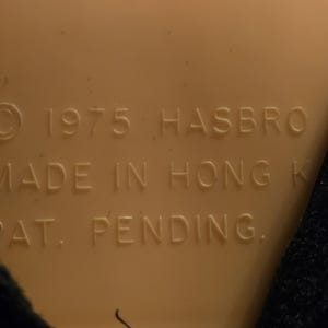 Peut inclure: Surface en plastique beige avec le texte "&copy; 1975 HASBRO MADE IN HONG K PAT. PENDING." en marron fonc&eacute;. Un tissu noir est visible sur le c&ocirc;t&eacute; droit de l'image.