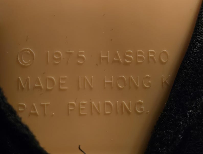 Peut inclure: Surface en plastique beige avec le texte "&copy; 1975 HASBRO MADE IN HONG K PAT. PENDING." en marron fonc&eacute;. Un tissu noir est visible sur le c&ocirc;t&eacute; droit de l'image.