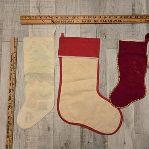 May include: Three vintage Christmas stockings. One is white with a red cuff and a printed design. Another is white with a red cuff and a plain design. The third is red velvet with a gold trim.