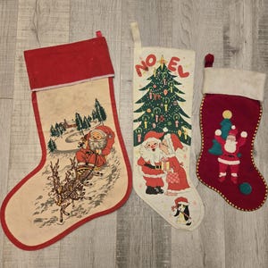 May include: Three vintage Christmas stockings with different designs. One stocking features a red background with a gold trim and a Santa Claus illustration. Another stocking has a white background with a red trim and a Santa Claus and reindeer illustration. The third stocking has a white background with a green Christmas tree and the word "NOEL" in red.