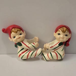 May include: Two ceramic elf figurines. Each elf wears a red hat and a cream-colored onesie with red and green stripes. The elves have smiling faces and are posed in a sitting position.