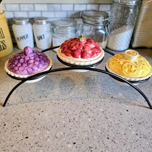 May include: A decorative serving piece with three ceramic pie-shaped dishes, each with a different fruit design. The dishes are purple, red, and yellow, and sit on a black metal stand. The background includes kitchen items like jars and a yellow container.