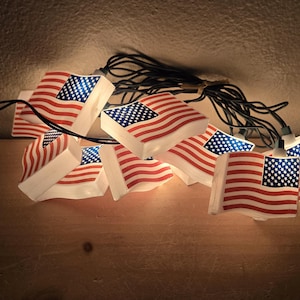 May include: A string of decorative lights featuring multiple small, illuminated plastic covers shaped like the American flag. Each flag cover displays the classic red, white, and blue design with stars and stripes, connected by a black electrical cord.