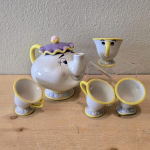 May include: A ceramic teapot with a purple ruffled lid and yellow handle, accompanied by three small matching teacups. The teapot features a painted face with closed eyes, while one teacup displays a painted face with open eyes and a yellow handle.