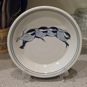 May include: A white ceramic plate with a blue rim and a painted design of five birds perched on a branch. The birds are in shades of blue and gray. The plate is displayed on a clear plastic stand.