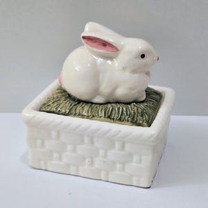 May include: A white ceramic bunny figurine with pink inner ears and a pink tail sits atop a square basket-shaped container. The container has a woven texture and is filled with green faux grass.