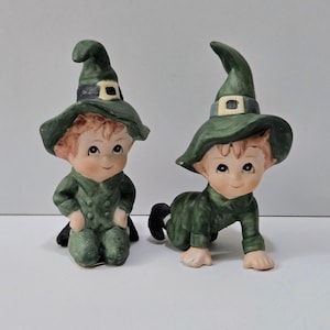 May include: Two ceramic figurines of small figures wearing green witch hats and outfits. One is kneeling, the other is crawling. The hats have a black band with a gold buckle. The figures have rosy cheeks and are smiling.
