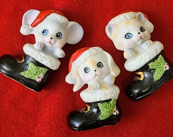May include: Three ceramic Christmas ornaments, each depicting a different animal wearing a Santa hat and sitting in a black boot with holly leaves. The animals are a mouse, a cat, and a dog.