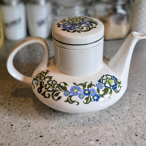 May include: White teapot with a floral design featuring blue and green flowers. The teapot has a lid with a matching floral design.