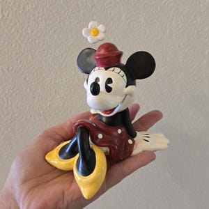 Puede incluir: Una figura de Minnie Mouse, con una falda roja de lunares blancos, una blusa negra y zapatos amarillos. La figura tiene un sombrero rojo y una flor blanca encima de la cabeza. La figura est&aacute; sentada y sonriendo.