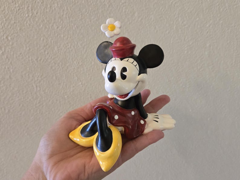 Puede incluir: Una figura de Minnie Mouse, con una falda roja de lunares blancos, una blusa negra y zapatos amarillos. La figura tiene un sombrero rojo y una flor blanca encima de la cabeza. La figura est&aacute; sentada y sonriendo.