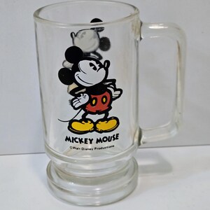May include: A clear glass mug featuring a graphic of Mickey Mouse wearing red shorts and yellow shoes. The text MICKEY MOUSE is printed in black below the character, followed by the smaller text Walt Disney Productions.