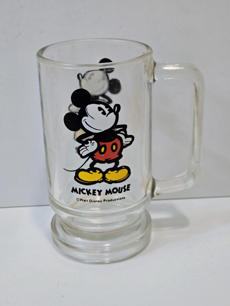 May include: A clear glass mug featuring a graphic of Mickey Mouse wearing red shorts and yellow shoes. The text MICKEY MOUSE is printed in black below the character, followed by the smaller text Walt Disney Productions.