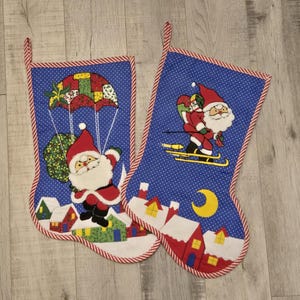 May include: Two Christmas stockings with blue backgrounds and white polka dots. One stocking features Santa parachuting, the other shows Santa skiing. Both have red and white striped trim and a village scene at the bottom.