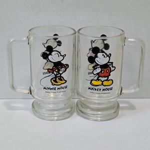 May include: Two clear glass mugs with handles and pedestal bases. One mug features a graphic of Minnie Mouse with the text Minnie Mouse and Walt Disney Productions. The other mug features Mickey Mouse with the text Mickey Mouse and Walt Disney Productions.