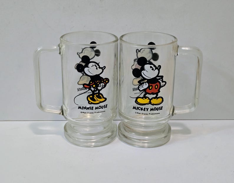 May include: Two clear glass mugs with handles and pedestal bases. One mug features a graphic of Minnie Mouse with the text Minnie Mouse and Walt Disney Productions. The other mug features Mickey Mouse with the text Mickey Mouse and Walt Disney Productions.