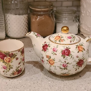 May include: A white teapot and mug with a floral pattern featuring red, yellow, and pink roses. The teapot has a gold lid and handle. The mug has a gold rim.