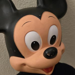 Peut inclure: Gros plan d'un masque de Mickey Mouse. Le masque pr&eacute;sente une t&ecirc;te et des oreilles noires, un visage couleur p&ecirc;che, des yeux blancs et un nez noir. Le masque est pos&eacute; sur un fond neutre et porte une chemise noire.