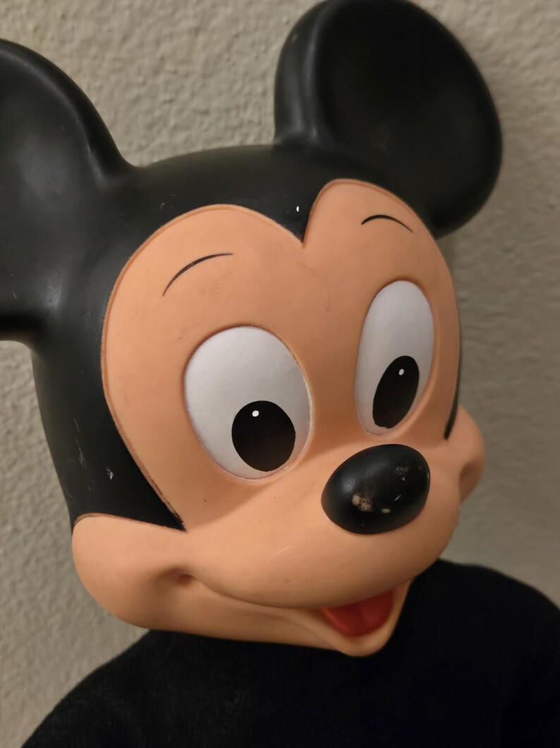 Peut inclure: Gros plan d'un masque de Mickey Mouse. Le masque pr&eacute;sente une t&ecirc;te et des oreilles noires, un visage couleur p&ecirc;che, des yeux blancs et un nez noir. Le masque est pos&eacute; sur un fond neutre et porte une chemise noire.