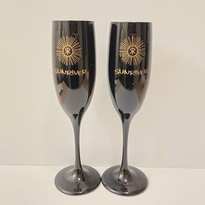 May include: Two black champagne flutes with gold-colored text and a sunburst design. The text on the flutes reads "SUNRIVER". The flutes have a slender stem and a wide base. The flutes are made of a shiny material.