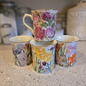 May include: Four ceramic mugs with floral designs. The mugs feature different floral patterns, including roses, irises, daffodils, and lilies. The mugs are stacked on top of each other.