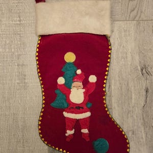 May include: Red velvet Christmas stocking with a white faux fur cuff. The stocking features an embroidered image of Santa Claus with a Christmas tree and a yellow star.