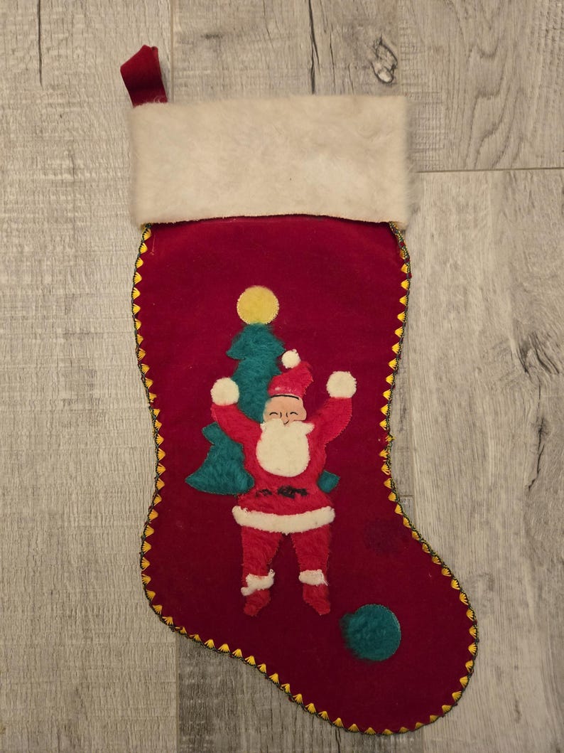 May include: Red velvet Christmas stocking with a white faux fur cuff. The stocking features an embroidered image of Santa Claus with a Christmas tree and a yellow star.