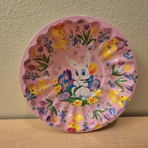 May include: A pink ceramic Easter egg dish with a scalloped edge. The dish is decorated with a white bunny holding a basket of eggs, surrounded by yellow chicks and flowers.