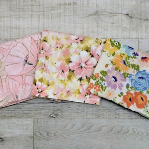 May include: Three folded floral fabric squares in pink, yellow, and white. The pink fabric features large, stylized flowers. The yellow fabric has a mix of pink and yellow flowers. The white fabric has blue, orange, and purple flowers.