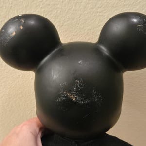 Peut inclure: T&ecirc;te noire de Mickey Mouse avec deux grandes oreilles rondes. La t&ecirc;te pr&eacute;sente des signes d'usure, avec des rayures et une d&eacute;coloration. Les oreilles sont lisses et arrondies, et la forme g&eacute;n&eacute;rale est embl&eacute;matique.