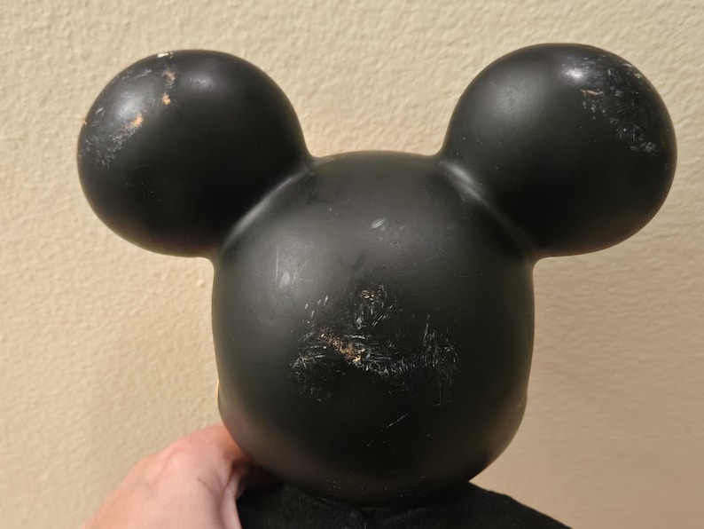Peut inclure: T&ecirc;te noire de Mickey Mouse avec deux grandes oreilles rondes. La t&ecirc;te pr&eacute;sente des signes d'usure, avec des rayures et une d&eacute;coloration. Les oreilles sont lisses et arrondies, et la forme g&eacute;n&eacute;rale est embl&eacute;matique.