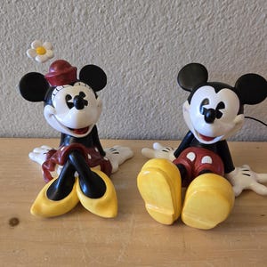 Puede incluir: Dos figuras de cer&aacute;mica de Mickey y Minnie Mouse. Minnie lleva un sombrero y una falda rojos, zapatos amarillos y una flor blanca. Mickey lleva pantalones cortos rojos y zapatos amarillos. Ambos tienen orejas negras y guantes blancos.