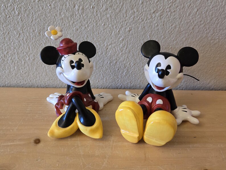 Puede incluir: Dos figuras de cer&aacute;mica de Mickey y Minnie Mouse. Minnie lleva un sombrero y una falda rojos, zapatos amarillos y una flor blanca. Mickey lleva pantalones cortos rojos y zapatos amarillos. Ambos tienen orejas negras y guantes blancos.