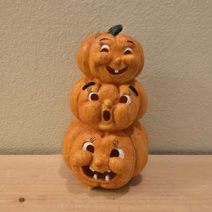 May include: A stack of three orange jack-o'-lantern pumpkins with carved faces. The top pumpkin has a smiling expression, the middle pumpkin has an open-mouthed surprised expression, and the bottom pumpkin has a wide, toothy, grinning expression.