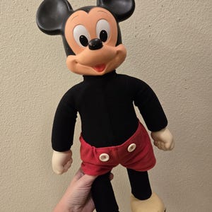 Peut inclure: Une poup&eacute;e en peluche vintage de Mickey Mouse. La poup&eacute;e a une t&ecirc;te noire avec de grandes oreilles, un visage beige et un col roul&eacute; noir. Elle porte un short rouge avec des boutons blancs, des jambes noires et des chaussures beiges.