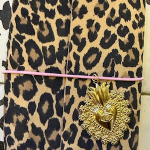 Peut inclure: Un journal avec une couverture à imprimé léopard, maintenu par un élastique rose. Un charm en forme de cœur doré avec un motif d'œil est suspendu à l'élastique. Le journal a des coins dorés.