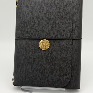 May include: A black leather-textured journal secured with a thin black elastic band. A circular gold-toned charm featuring an embossed eye design is attached to the centre of the elastic band. The journal has a pebbled surface and gold metal accents.
