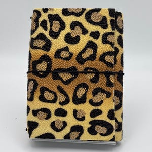 Può includere: Una copertina per taccuino rettangolare caratterizzata da un motivo leopardato testurizzato nei toni del nero, marrone chiaro e crema. La copertina è fissata da un sottile elastico nero avvolto orizzontalmente attorno al centro.