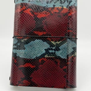 May include: A rectangular notebook cover featuring a textured snakeskin pattern in vibrant shades of red, deep burgundy, and light blue. The cover is secured by a thin black elastic band wrapped horizontally around the centre of the exterior.