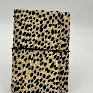 Puede incluir: Un cuaderno rectangular con una cubierta texturizada de color crema con un patrón repetitivo de manchas negras irregulares. Una fina banda elástica negra envuelve horizontalmente el centro de la cubierta, asegurada por pequeños ojales metálicos dorados.
