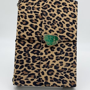 May include: A rectangular notebook with a tan and black leopard print cover. A thin black elastic cord wraps horizontally around the center, securing a single, irregular, polished green stone with a gold-toned metallic edge as a closure.