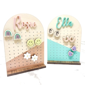 May include: Two wooden earring display stands with the names "Rosie" and "Ella" written on them. The stands are painted in a light wood colour and have a light blue and brown colour scheme. The stands have a variety of colourful earrings displayed on them.