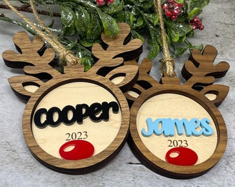 May include: Two wooden reindeer ornaments with the names "cooper" and "james" written on them. The ornaments are brown with a light wood center and have red noses. The year 2023 is written on each ornament.