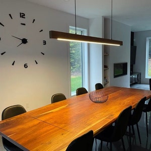 May include: A long wooden dining table with a live edge design, surrounded by black chairs. A modern light fixture hangs above the table, and a large clock is on the wall behind the table.