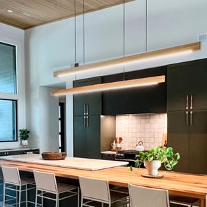 May include: Two linear pendant lights featuring long, rectangular light-colored wood housings with integrated LED strips suspended by thin black cables above a kitchen island with a natural wood countertop and white bar stools.