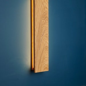 May include: A wooden wall sconce with a warm, glowing light emanating from a thin slit in the wood. The sconce is mounted on a blue wall.