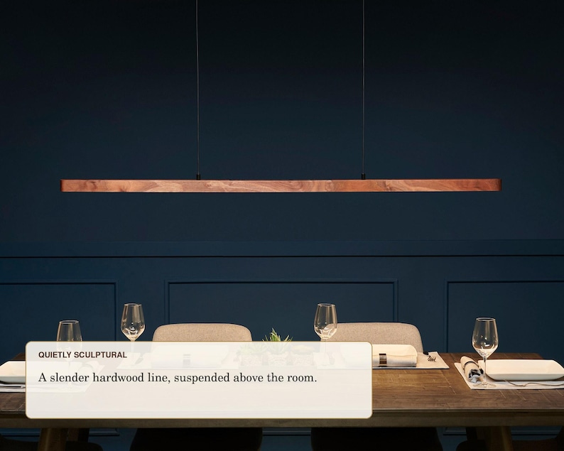 May include: A linear pendant light fixture featuring a long, slender piece of natural hardwood with a visible grain, suspended horizontally by two thin black cables. The text "QUIETLY SCULPTURAL A slender hardwood line, suspended above the room." is visible.